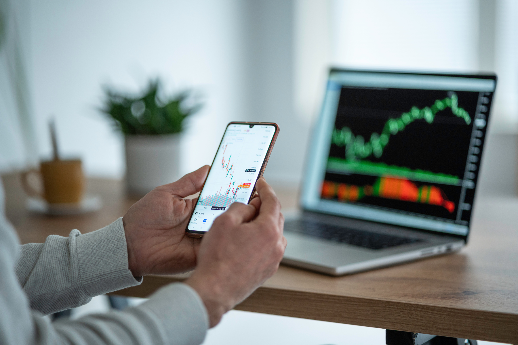 Person holding a mobile phone showing a trading chart, with a laptop in the background